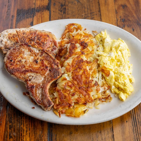 Two grilled pork chops, hash browns, and scrambled eggs on a white plate at Crazy Otto's Diner.