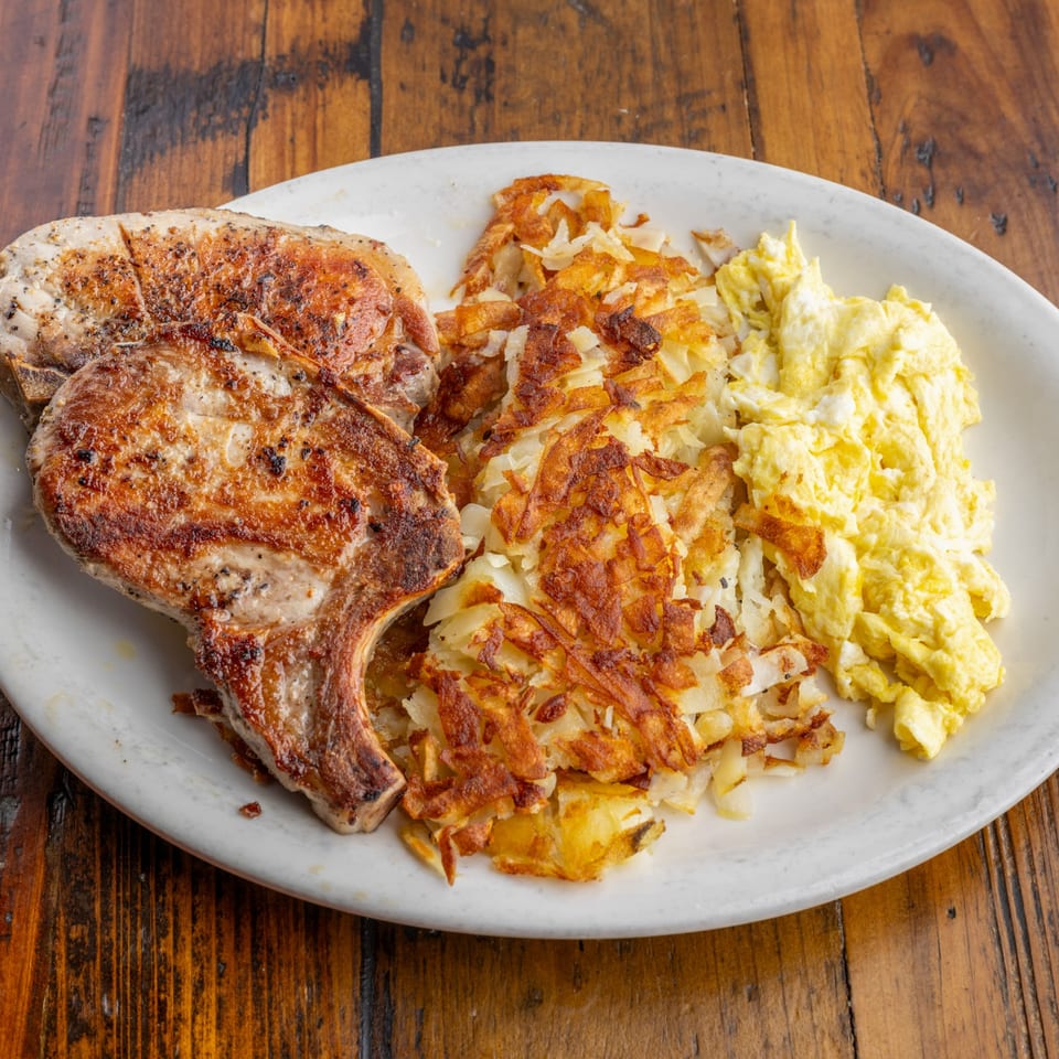 Two grilled pork chops, hash browns, and scrambled eggs on a white plate at Crazy Otto's Diner.
