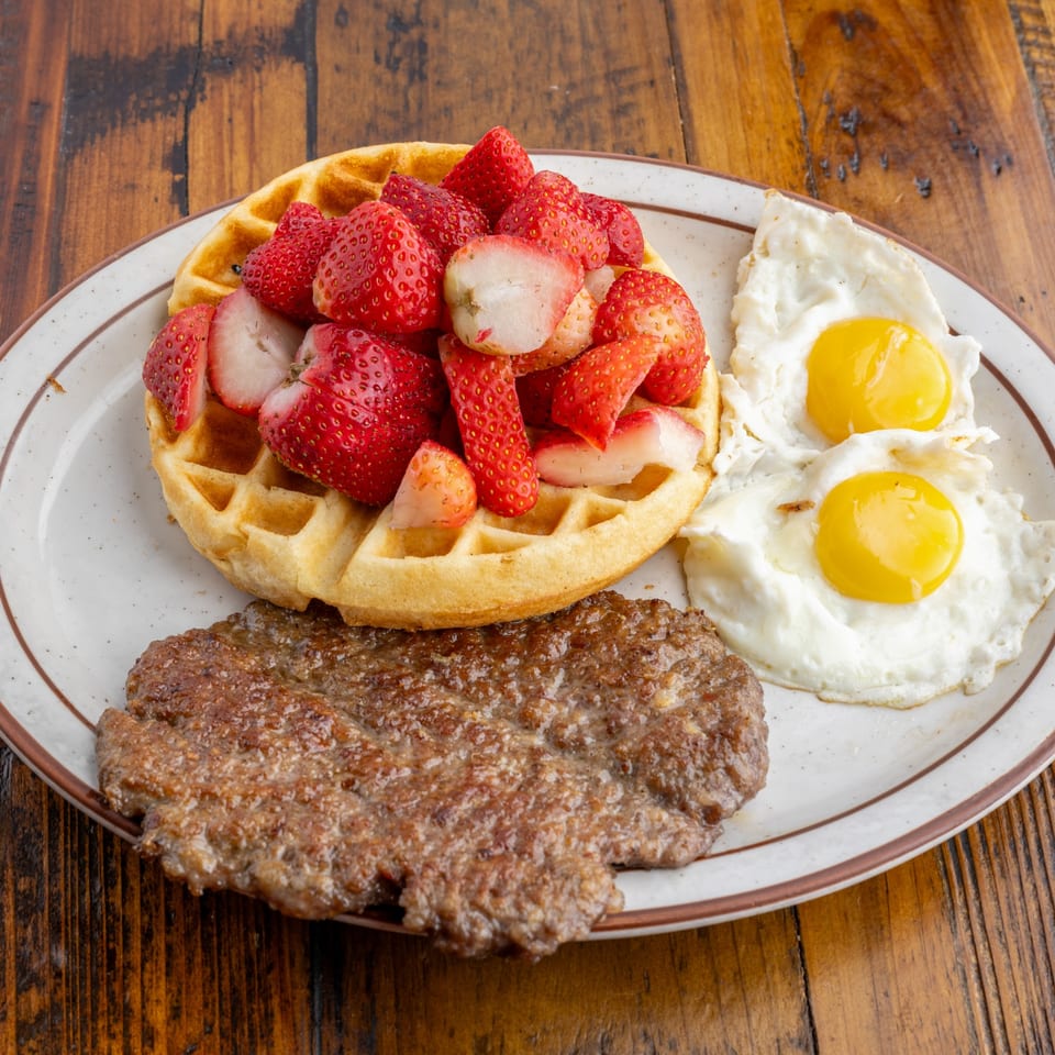 Crazy Otto's Diner breakfast with a waffle topped with strawberries, sausage patty, and two fried eggs.