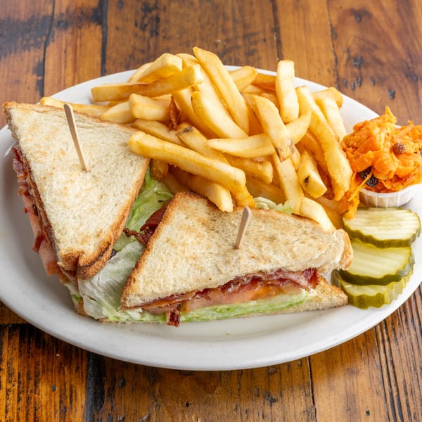 A diner club sandwich with crispy bacon, lettuce, and tomato, served with golden fries, coleslaw, and pickles.