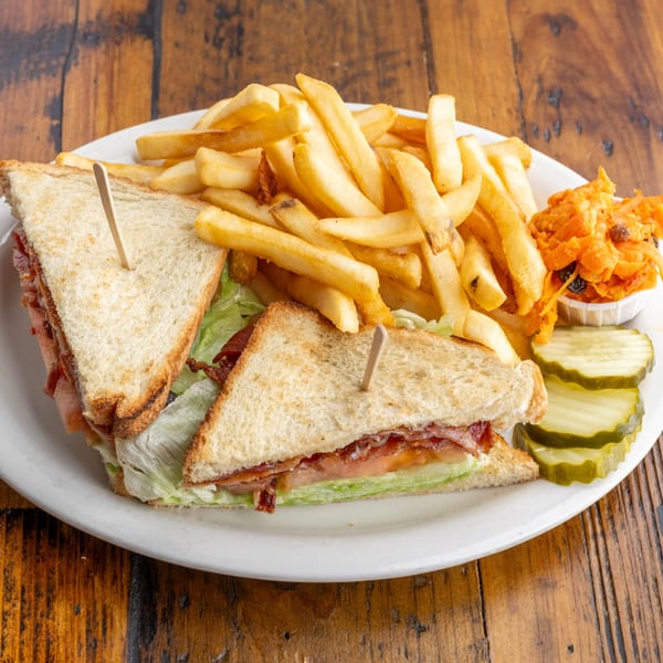 A Crazy Otto's Diner BLT club sandwich with fries, carrot salad, and pickles on a white plate.