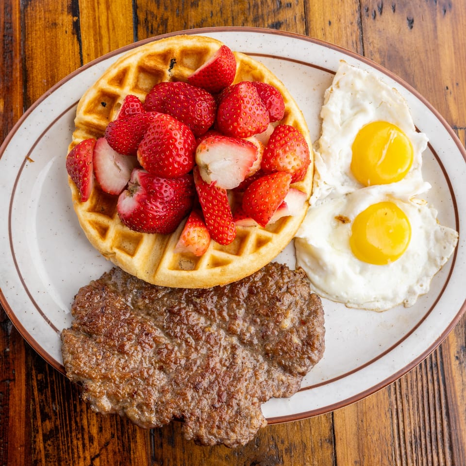 A Crazy Otto's Diner breakfast plate with a strawberry waffle, sausage patty, and two fried eggs.