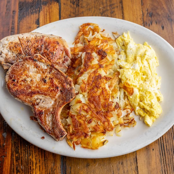 Two grilled pork chops, crispy hash browns, and fluffy scrambled eggs on a white plate.