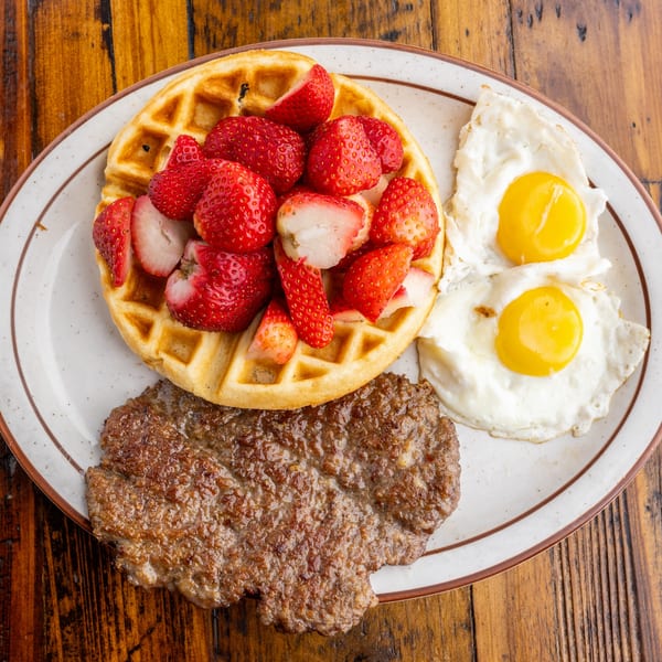 A diner breakfast with a strawberry-topped waffle, two fried eggs, and a sausage patty on a plate.