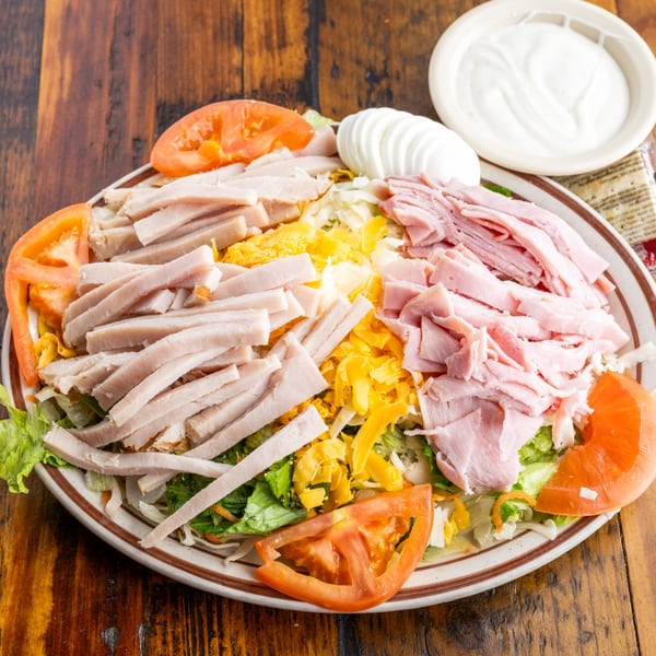 Chef salad with sliced turkey, ham, cheese, tomatoes, and hard-boiled egg on a wooden table.